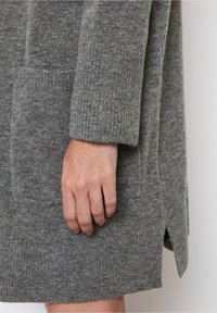 Šedý pletený cardigan s texturovaným povrchem, s žebrovanými manžetami, bočními kapsami a malým detailem s etiketou. Ruka odpočívá na látce.