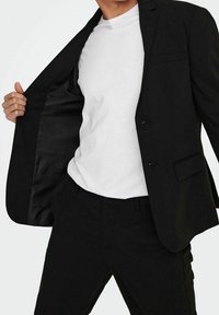 Czarna marynarka z satynowym wnętrzem, noszona na białej koszulce. Marynarka ma dopasowany fason, klapy w kształcie notched oraz jedną kieszeń na piersi.