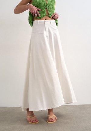 Person iført en hvid bukse med vide ben, grøn strikket crop top, beige flip-flops og sølvringe, stående på en grå gulv mod en ensfarvet væg.