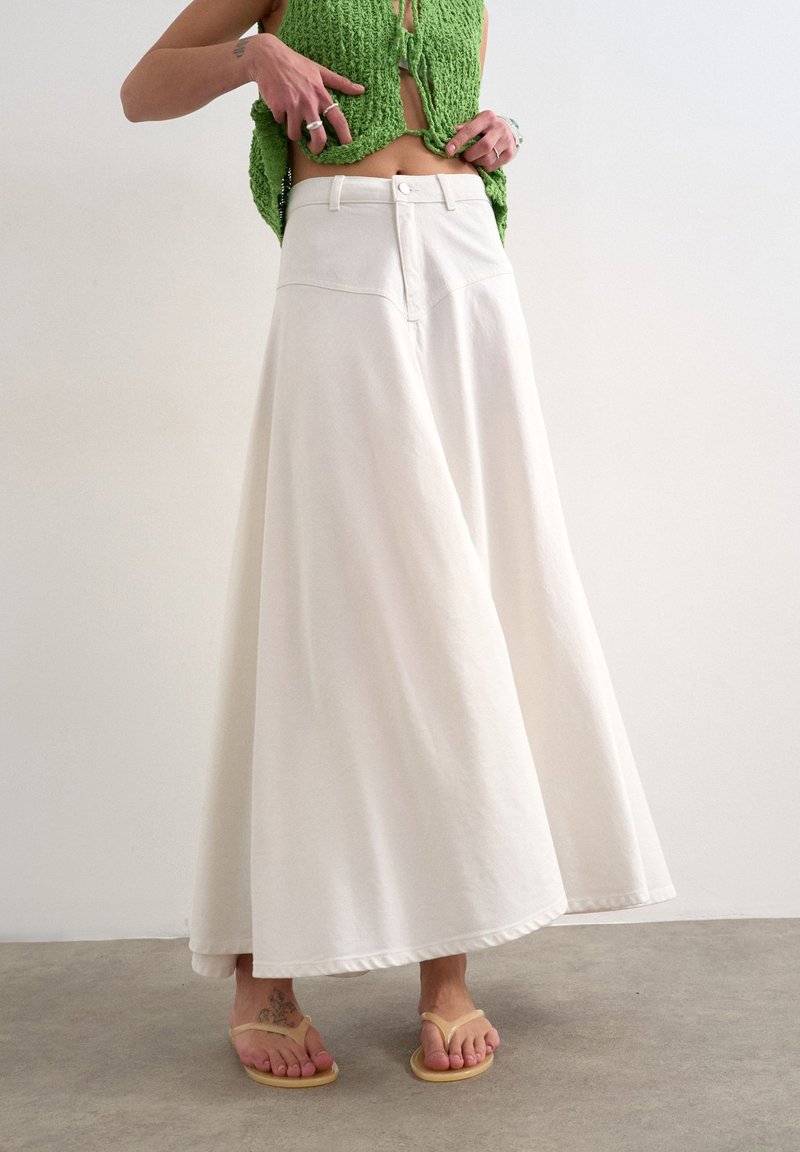 Personne portant une jupe blanche à jambes larges, un crop top en tricot vert, des tongs beiges et des bagues argentées, debout sur un sol gris contre un mur uni.