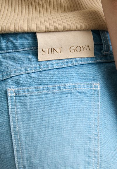 Blugi din denim albastru deschis cu o textură distinctă, având un eticheta din piele bej, embosată cu "STINE GOYA" pe talie.