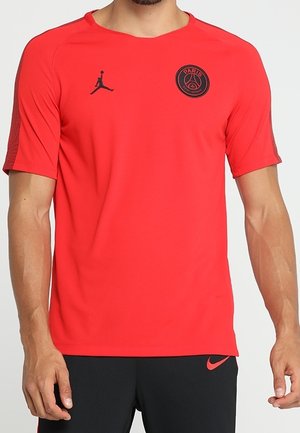 Mand iført en skarp rød kortærmet sportstrøje med sorte Paris Saint-Germain- og Jumpman-logoer, kombineret med sorte Nike-shorts.