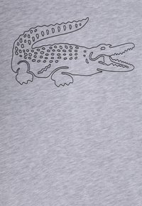 Szara koszulka z materiału z czarną konturową ilustracją krokodyla z detalami łusek i otwartą paszczą, które podkreślają prostotę designu.
