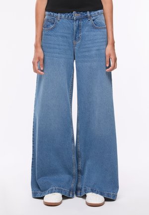 Persona che indossa jeans in denim azzurro chiaro a gamba larga e sneakers bianche con suola marrone su uno sfondo semplice.