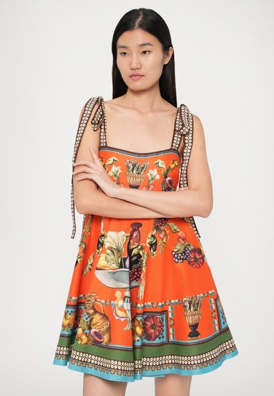 Femeie îmbrăcată într-o rochie fără mâneci de culoare portocalie, cu modele florale și de fructe, având curele legate la umăr, stând cu brațele încrucișate.