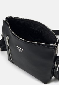 Borsa a tracolla sintetica nera con superficie testurizzata, hardware in tonalità argento, scomparto principale con zip e dettaglio del logo sul davanti.