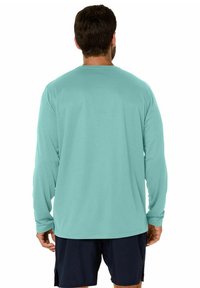 Camicia a maniche lunghe in teal chiaro, con una texture liscia e scollatura rotonda, indossata con pantaloni corti scuri, mostrando un design semplice sul retro.