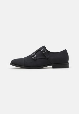 Chaussure habillée à boucle monastère en cuir noir avec doubles boucles argentées, bout pointu et petit talon sur fond blanc.