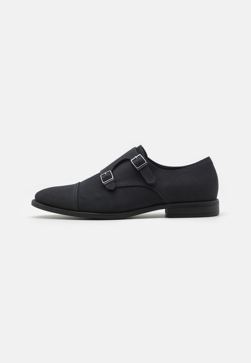 Chaussure habillée à boucle monastère en cuir noir avec doubles boucles argentées, bout pointu et petit talon sur fond blanc.