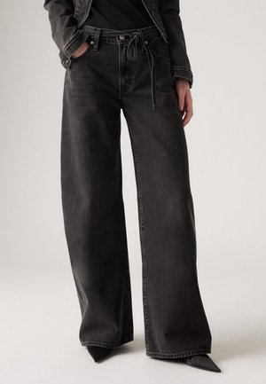 Personne portant un jean en denim noir à jambes larges avec une taille à cordon, mains dans les poches, assorti à des chaussures noires pointues et une veste noire.