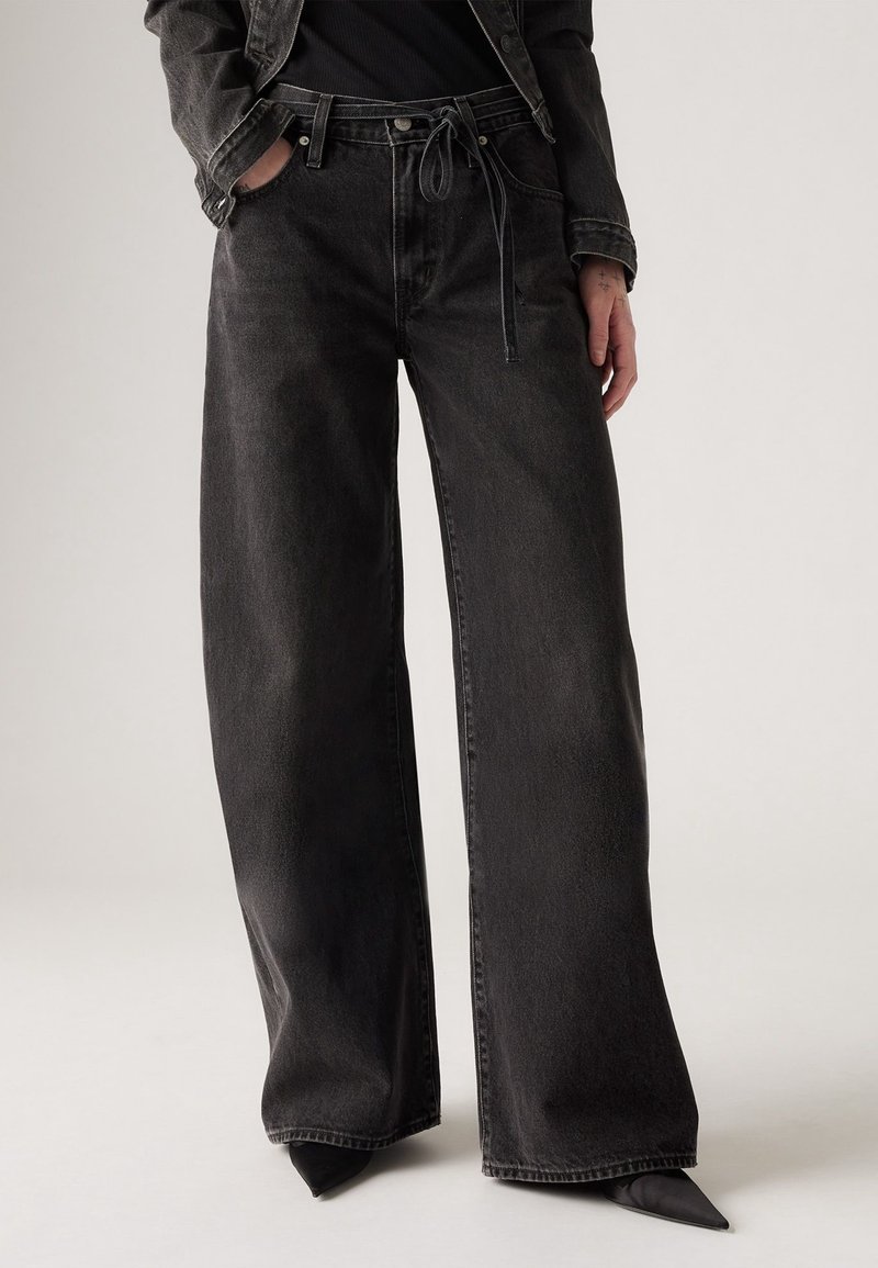 Personne portant un jean en denim noir à jambes larges avec une taille à cordon, mains dans les poches, assorti à des chaussures noires pointues et une veste noire.