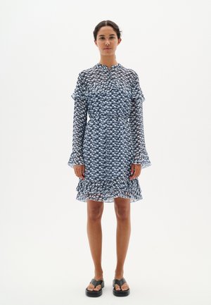 Leichtes, blau gemustertes Kleid mit langen Ärmeln, Rüschensaum und gestuftem Saum. Besitzt einen Kragen und einen lässigen Schnitt, kombiniert mit schwarzen Sandalen.