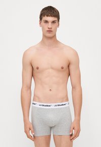 Graue Boxershorts mit einem weißen elastischen Bund, auf dem "JJ Studios" in Schwarz gedruckt ist. Der Stoff scheint weich und eng anliegend zu sein.