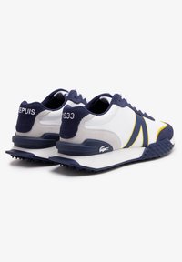 Zapatillas deportivas para hombre con parte superior sintética blanca, acentos en azul marino y rayas amarillas. Cuentan con una suela de goma texturizada y un collar acolchado.