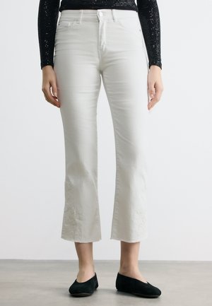 Λευκά τζιν καμπάνα με κομμένο μήκος και διακριτική κεντημένη λουλουδάτη λεπτομέρεια κοντά στο τελείωμα, φορεμένα με μαύρα μπαλαρίνες και μαύρο μακρυμάνικο τοπ.