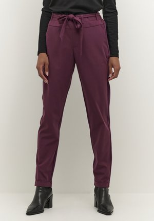 Personne portant un pantalon prune taille haute avec ceinture nouée, un haut noir à manches longues et des bottines noires à talons, debout sur un fond uni.
