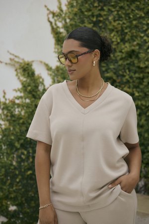 Top beige a scollatura a V con maniche corte, realizzato in materiale morbido. Presenta dettagli di cucitura e una vestibilità comoda. Abbinato a pantaloni e accessori coordinati.