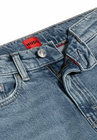 Blå denimjeans med en röd "HUGO"-etikett, silverknapp och dragkedjestängning med en sliten textur och klassisk femficksdesign.