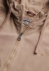 Beżowa bluza z kapturem na suwak z sznurkami, częściowo rozpięta, ukazująca metkę "Superdry Outerwear" wewnątrz kołnierza.