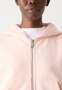 Felpa con zip rosa chiaro, realizzata in tessuto morbido, dotata di tasca frontale, zip argentata e cappuccio integrato. Design semplice e funzionale.