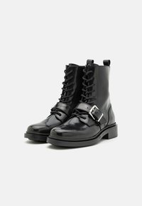 Botas de tobillo de charol negro con cordones frontales, hebillas plateadas y punta redondeada. Presentan un tacón bajo tipo bloque y una pestaña trasera.