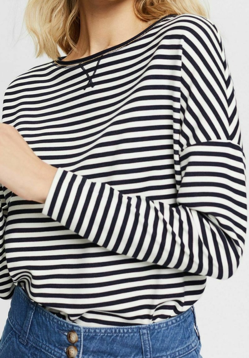 Maglietta a maniche lunghe a righe bianche e nere con scollo rotondo, caratterizzata da una vestibilità rilassata e dettagli di cuciture a contrasto. Abbinata a una gonna di denim.