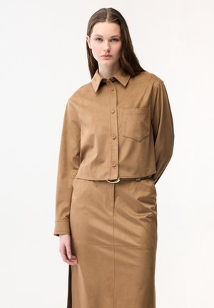 Jeune femme debout, portant une chemise courte boutonnée couleur beige avec une poche et une jupe taille haute assortie fendue, fond neutre.