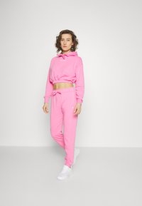 Cappuccio corto rosa con coulisse, orlo a coste e pantaloni abbinati. Texture liscia con logo sui top e pantaloni. Sneakers bianche.