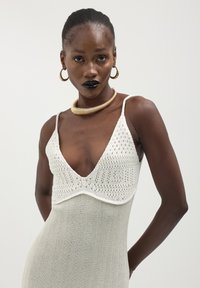Abito in maglia con scollatura a V profonda, corpetto in crochet bianco e gonna grigia testurizzata, dotato di spalline sottili e accessori minimalisti in oro.