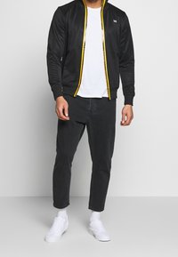Sort zip-up bomberjakke med gule detaljer, parret med en hvid t-shirt og sorte denimjeans. Har lange ærmer og en tætsiddende design.