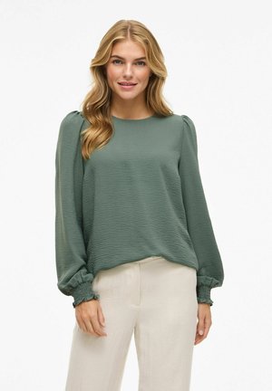 Blouse verte à manches longues en tissu texturé. Elle possède des épaules bouffantes et des poignets froncés. Associée à un pantalon ample de couleur claire.