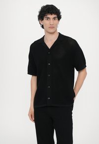 Juodas trumpomis rankovėmis marškinėliai su mygtukais, pagaminti iš tekstūruotos trikotažo medžiagos. Yra klasikinis apykaklė ir penki priekiniai mygtukai.