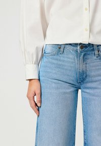 Jasnoniebieskie dżinsy z wysokim stanem, charakteryzujące się lekko zwężającą się nogawką i subtelnym wypłowieniem. W zestawieniu z białą koszulą zapinaną na guziki.