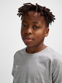 Zbliżenie młodej osoby czarnoskórej z krótkimi skręconymi włosami, ubranej w jasnoszarą koszulkę, spokojnie patrzącej w kamerę na jednolitym tle.