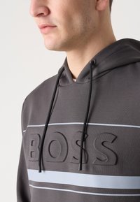 Felpa grigia con un logo "BOSS" testurizzato, caratterizzata da una finitura liscia, cordini neri e una striscia orizzontale grigia chiara sul petto.