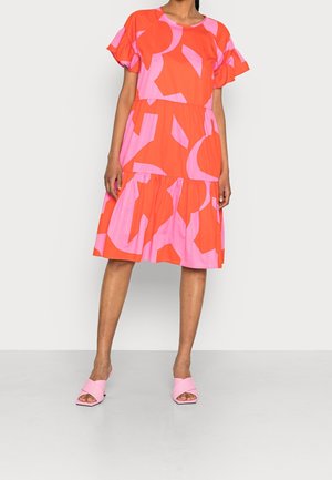 Femme portant une robe à manches courtes, arrivant au genou, avec des motifs abstraits rouges et roses, et des sandales à talons ouverts roses, se tenant devant un fond uni.