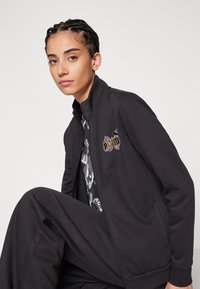 Giacca nera con zip realizzata in tessuto morbido, caratterizzata da un design floreale ricamato sul petto. Indossata sopra uno strato di abbigliamento con motivo.