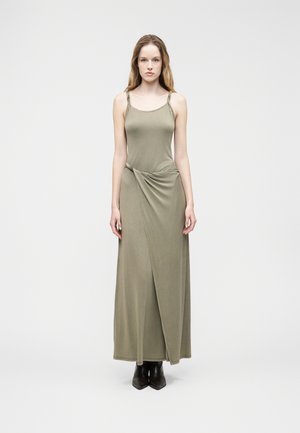 Kvindelig model stående iført en ærmeløs olivengrøn omlott-maxikjole med snoede stropper og sorte ankelstøvler på ensfarvet baggrund.