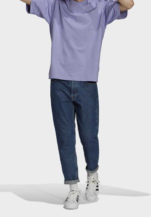 Persona vistiendo una camiseta morada, jeans azules remangados, calcetines blancos y zapatillas blancas con rayas negras, de pie con los brazos levantados.