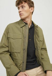 Olivgrön lätt jacka med dragkedja fram, två bröstfickor och tryckknappar, lager över en svart polo skjorta.