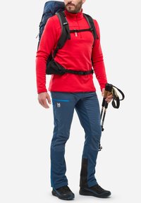 Homme portant une chemise à manches longues rouge, un pantalon de randonnée bleu, des chaussures de randonnée noires, portant un grand sac à dos bleu, tenant deux bâtons de randonnée.