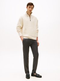 Krämfärgad zip-up polotröja med ett strukturerat geometriskt mönster, ficka framtill; kombinerad med smala mörkgrå byxor och svarta skor.