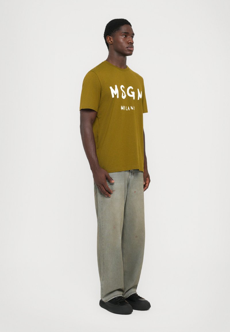 Olivgrön bomull t-shirt med vit "MSGN MILAN" tryck, tillsammans med ljusgrå vida jeans och svarta sneakers.