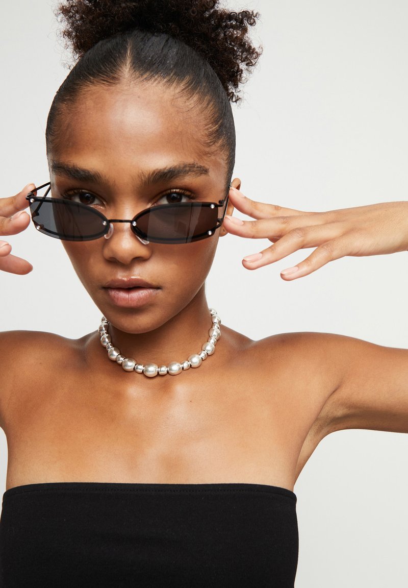 Gafas de sol ovaladas negras con lentes transparentes, choker de cuentas plateadas y una blusa negra sin tirantes. Textura suave, diseño moderno.