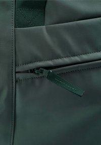 Grüner Stoffrucksack-Bereich mit einer Reißverschlusstasche und passendem grünen Reißverschlussgriff; glatte Textur mit verstärkten Nähten.