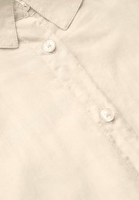 Beige katoenen shirt met een puntkraag, zichtbare stiksels en ronde witte knopen. Gladde textuur met een subtiele glans.