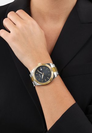 Gold- und silberfarbene Edelstahl-Uhr mit schwarzem Zifferblatt, geriffelter Lünette, goldenen Akzenten und Datumsanzeige. Wird am Handgelenk mit einem dunklen Outfit getragen.