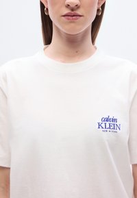Άσπρο βαμβακερό T-shirt με στρογγυλή λαιμόκοψη, κοντά μανίκια και μοβ κεντητό λογότυπο "Calvin Klein New York" στο αριστερό μέρος του στήθους.