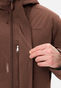 Veste imperméable marron avec une fermeture éclair à l'avant et une capuche. Présente une texture lisse, des coutures visibles et un logo sur la poitrine.