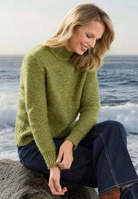 Frau in grünem Strickpullover und blauen Jeans, die auf einem Felsen am Ozean sitzt, lächelt und passt ihren Ärmel an.